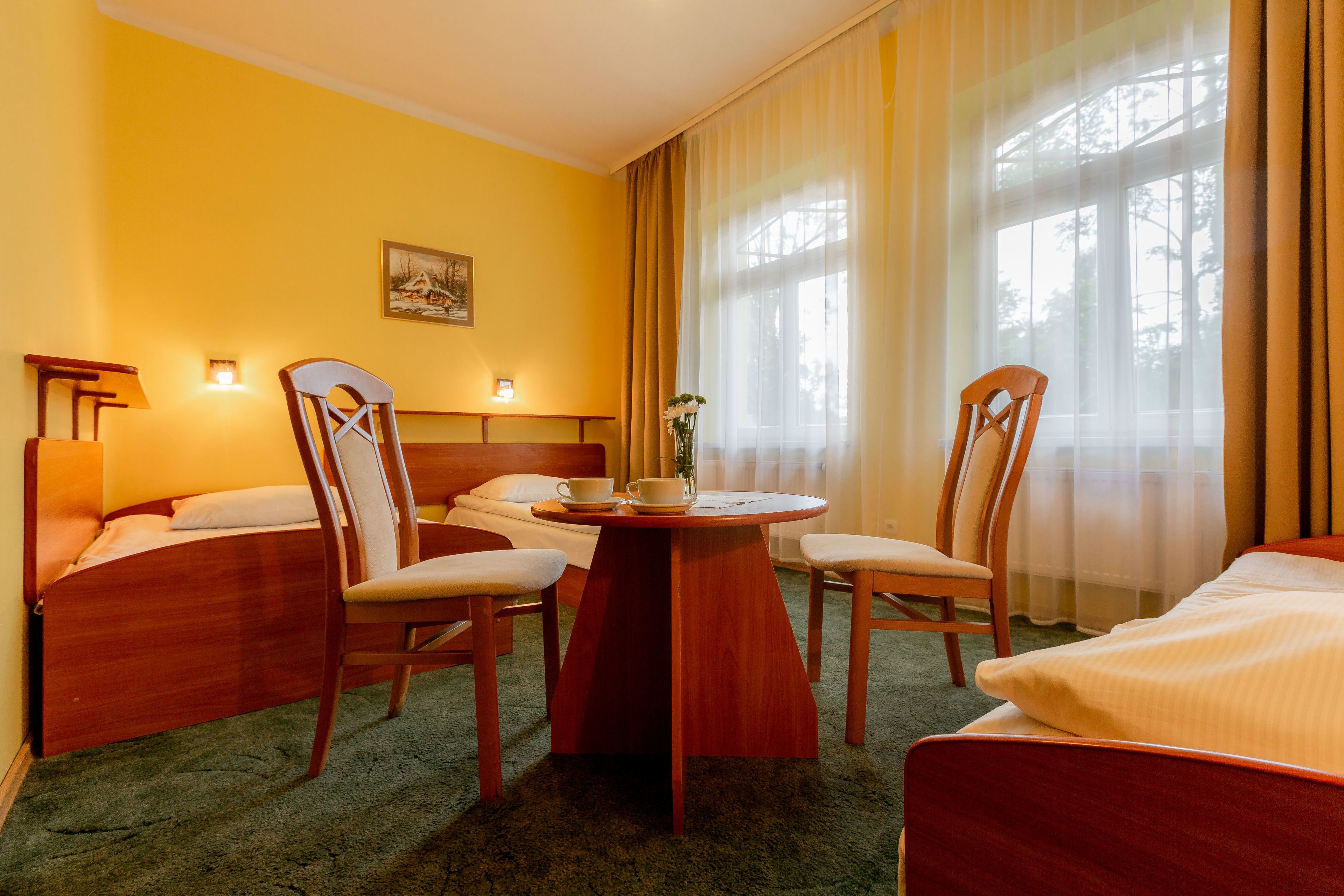 A hotel room with a bed, two chairs, a small table with a cup and saucer, and a painting on the wall.