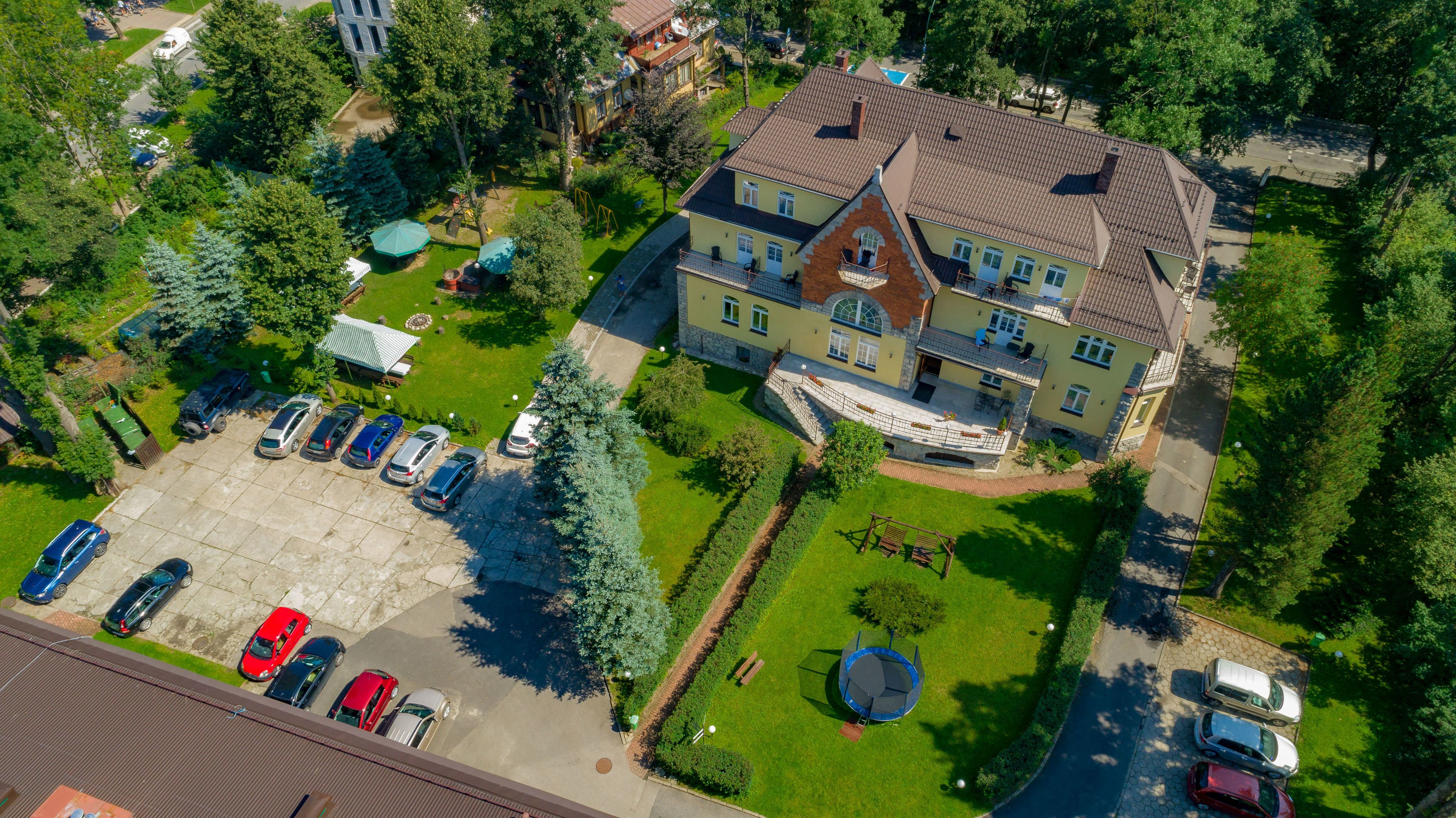 An aerial view of a large, multi-story house with a spacious front yard, a driveway with parked cars, and a small playground area.