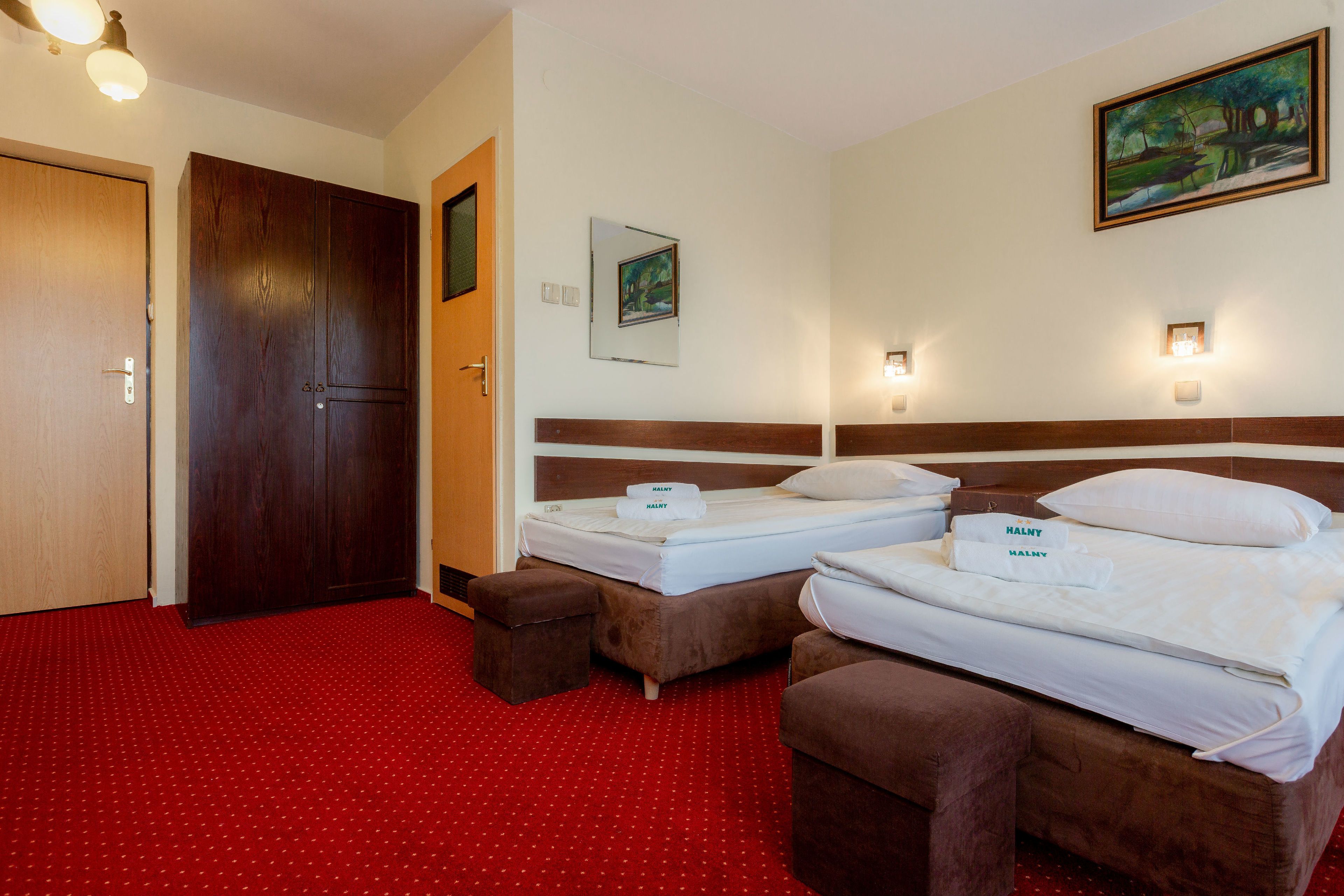A hotel room with two beds, a red carpet, a wooden wardrobe, a painting, and wall sconces.