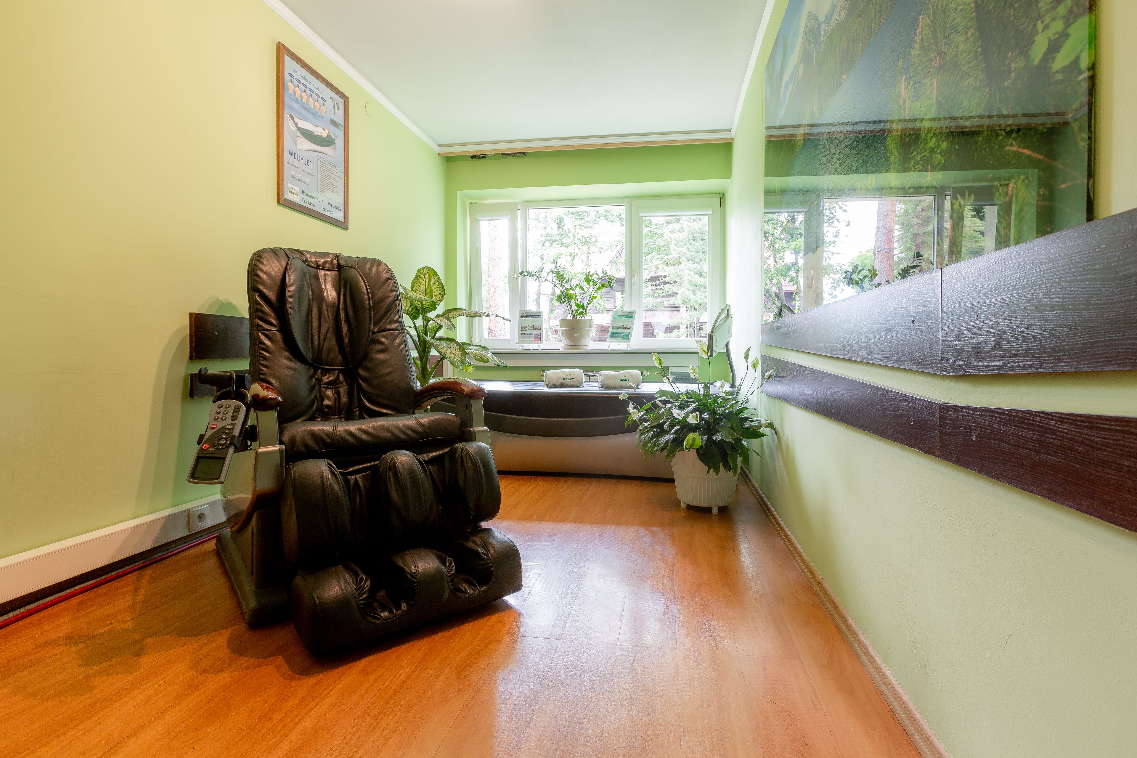 A massage chair in a room with a wooden floor, a window with plants, and a framed picture on the wall.