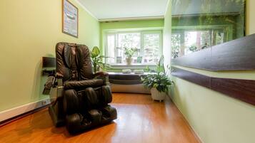 A massage chair in a room with a wooden floor, a window with plants, and a framed picture on the wall.