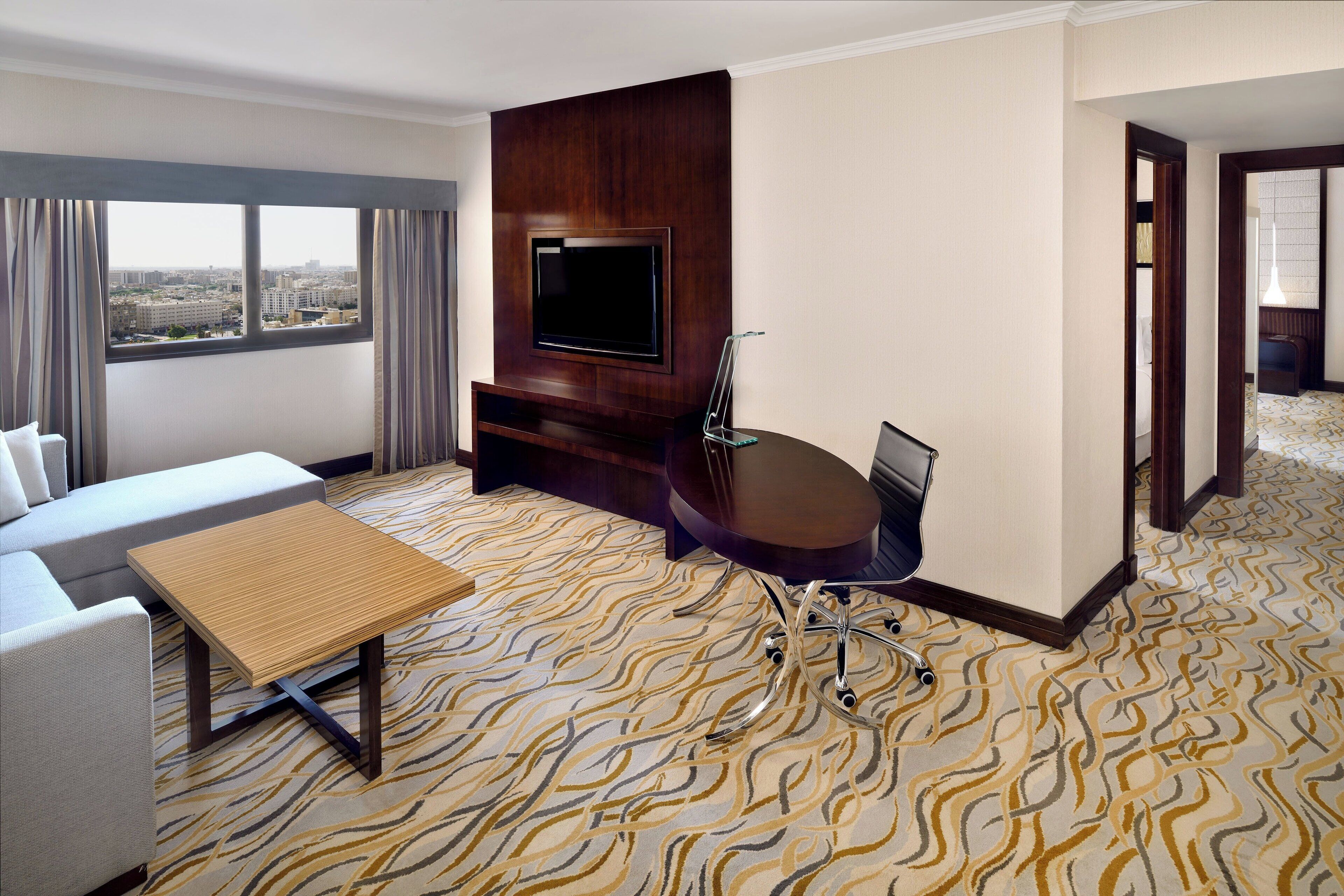 A hotel room with a sofa, a small table, a TV mounted on a wooden panel, a chair, and a patterned carpet.