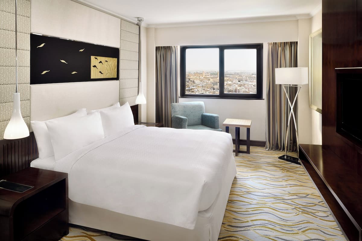 A hotel room with a large bed, a small table, a chair, a TV, and a window with a city view.