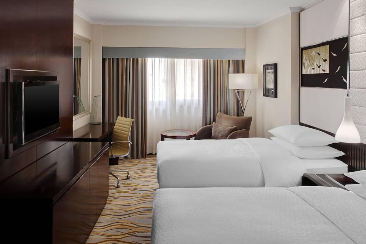 A hotel room with two beds, a desk, a chair, a TV, and a window with curtains.
