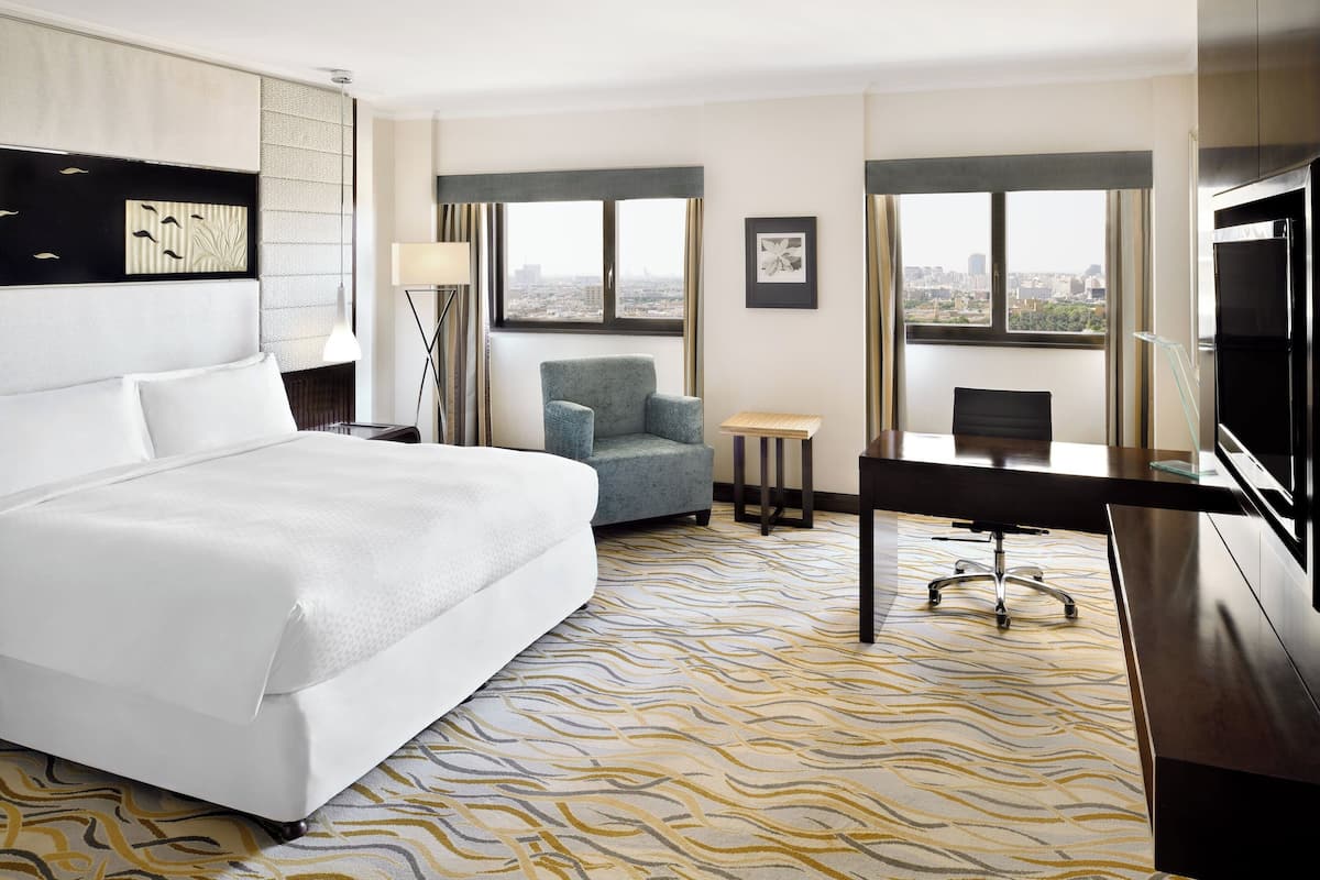 A hotel room with a large bed, a desk, a chair, and a TV. There are windows with a city view.