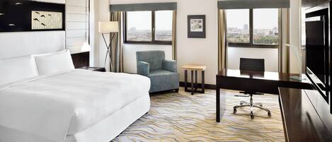 A hotel room with a large bed, a desk, a chair, and a TV. There are windows with a city view.