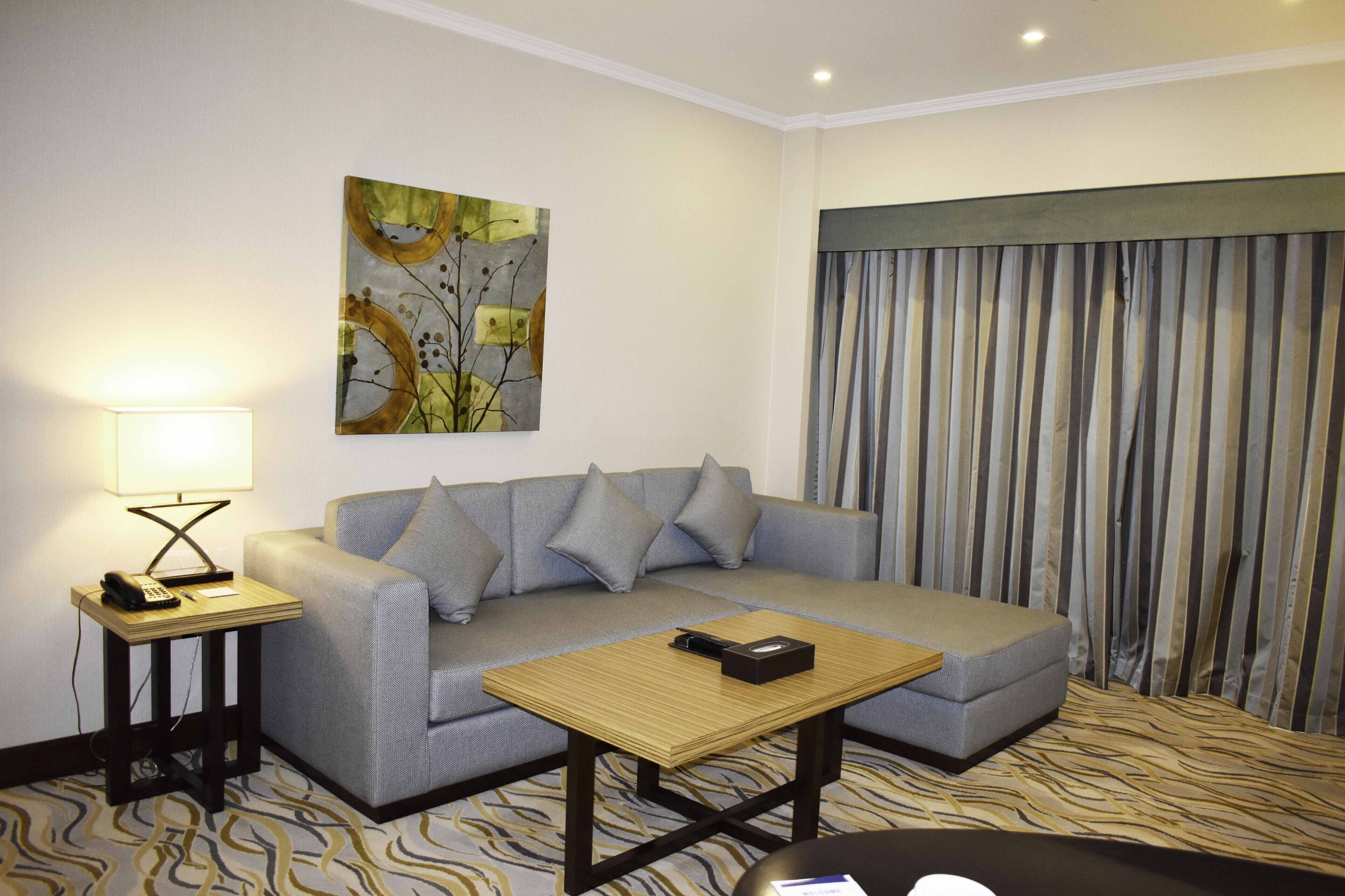 A modern hotel room with a grey sofa, a wooden coffee table, a side table with a lamp, and a large abstract painting on the wall.