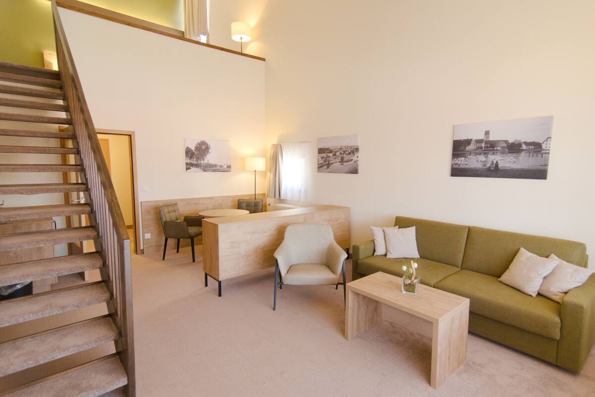 A modern living room with a staircase, a green sofa, a wooden coffee table, and a desk with chairs.