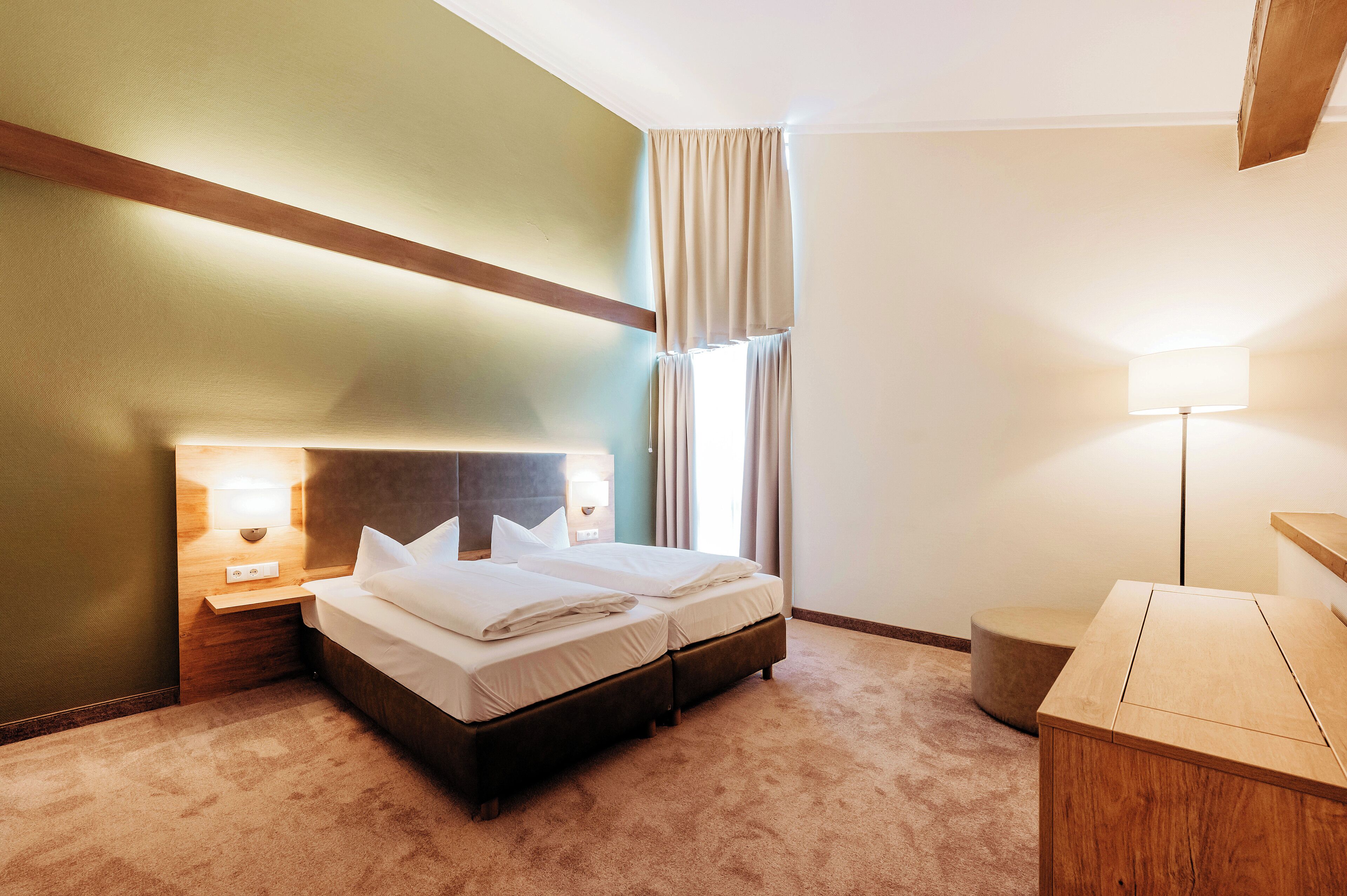 A hotel room with a double bed, bedside lamps, a wooden nightstand, a small wooden table, and a window with curtains.