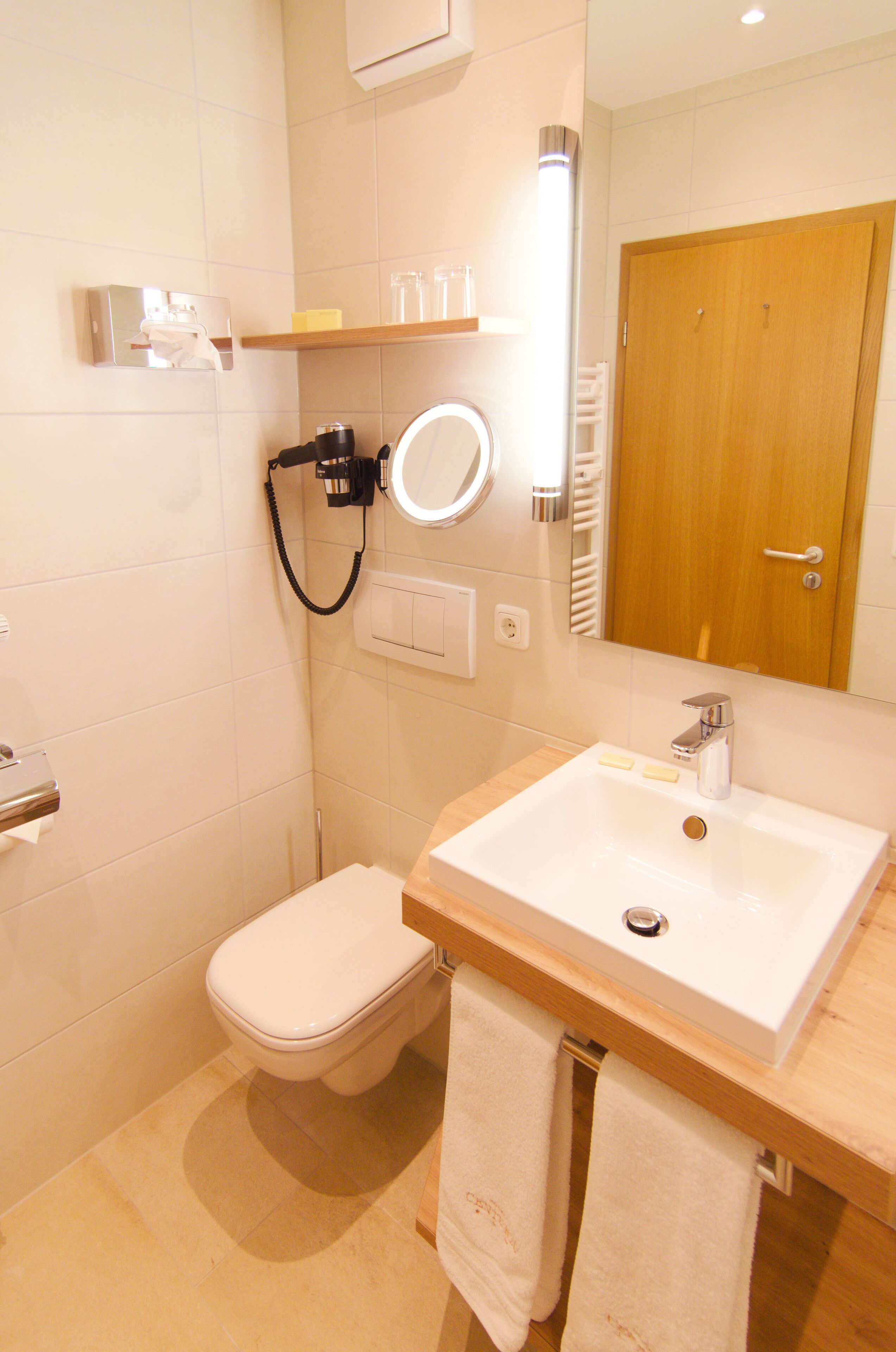A bathroom with a white sink, mirror, and toilet. There is a towel rack with two towels, a soap dispenser, and a hand dryer.