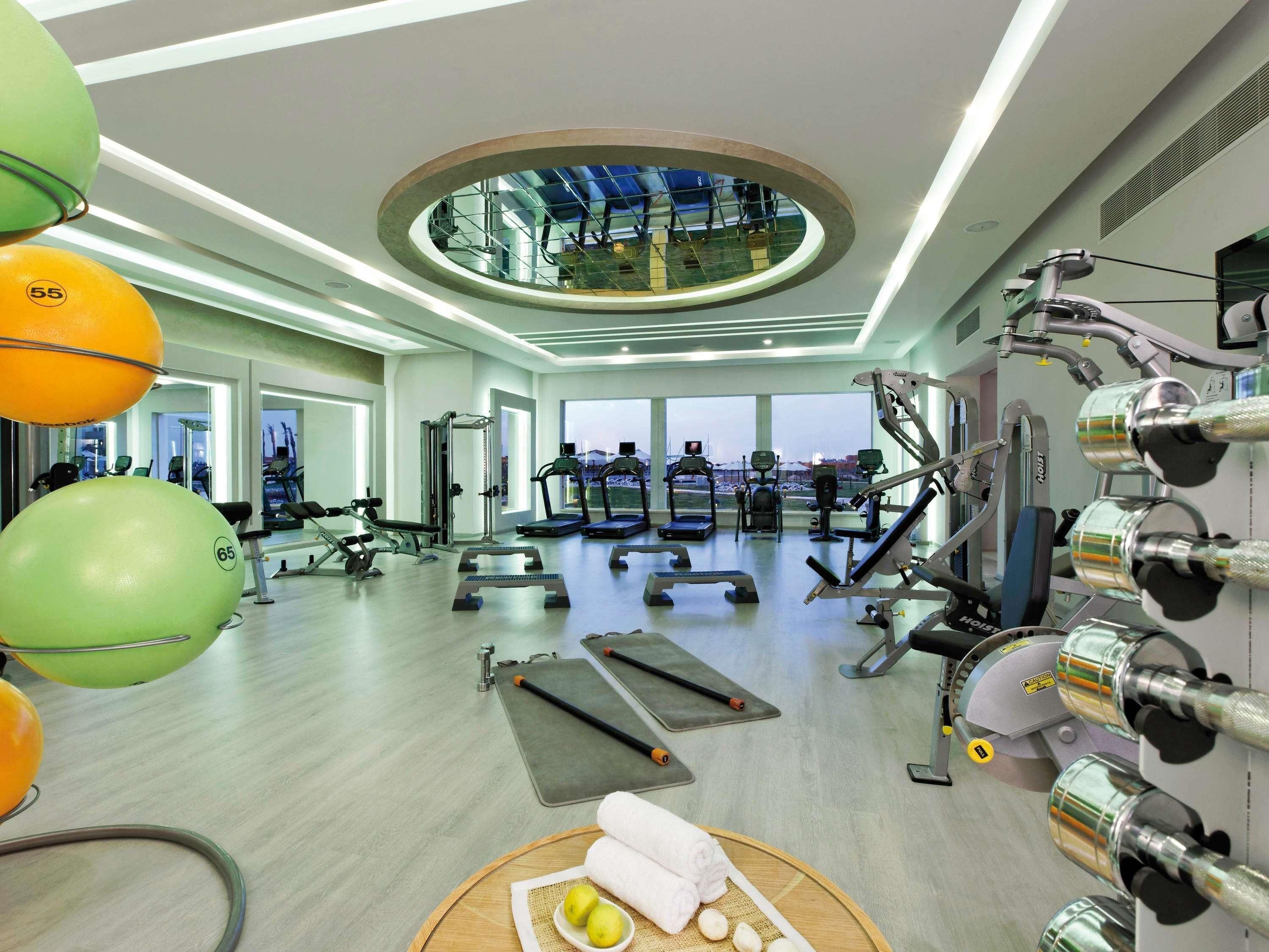 A modern gym with various exercise equipment, large windows, and a view of the cityscape.