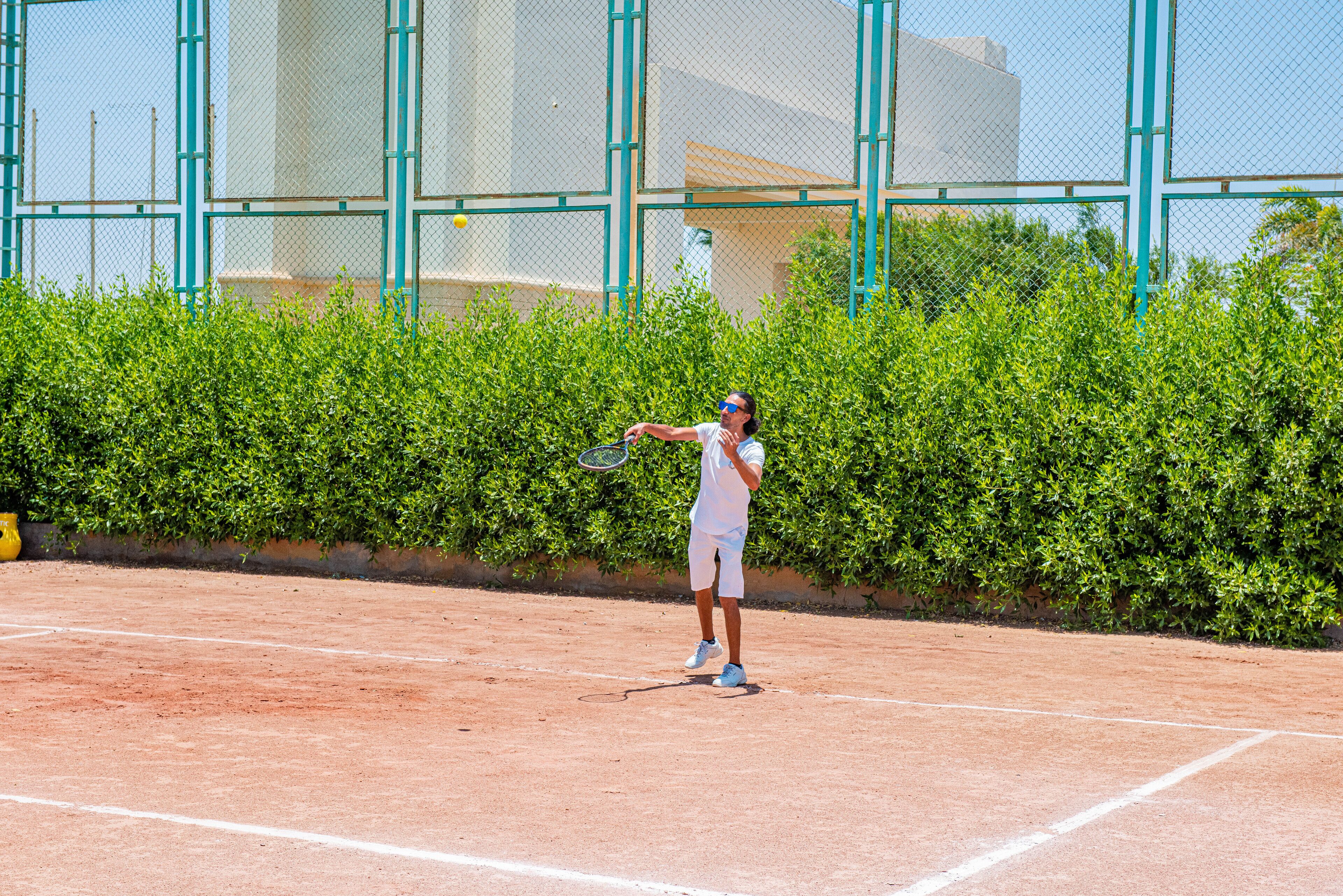 A tennis court with a player in white attire, a fence, and a hedge in the background.