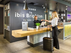 Bancada de recepção do ibis Brighton City Centre com design moderno, um laptop, uma planta e uma placa de boas-vindas.