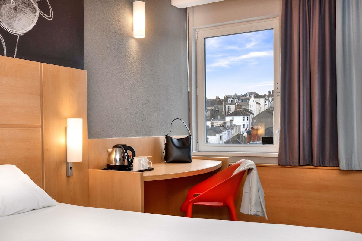 Habitación de hotel con cama, escritorio con hervidor y taza, una silla, ventana con cortinas y vista a la ciudad.