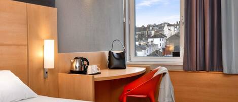 Une chambre d’hôtel comprenant un lit, un bureau avec une bouilloire et une tasse, une chaise, une fenêtre avec des rideaux et une vue sur la ville.