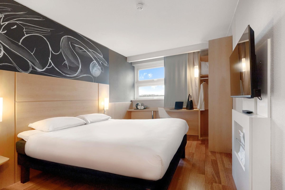 Una habitación de hotel moderna con una cama grande, un escritorio con una computadora portátil, un televisor de pantalla plana y una ventana con vista a la ciudad.