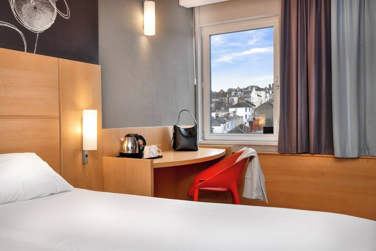 Habitación de hotel con cama, escritorio con silla, cafetera, bolso y ventana con cortinas.