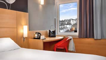 Habitación de hotel con cama, escritorio con silla, cafetera, bolso y ventana con cortinas.