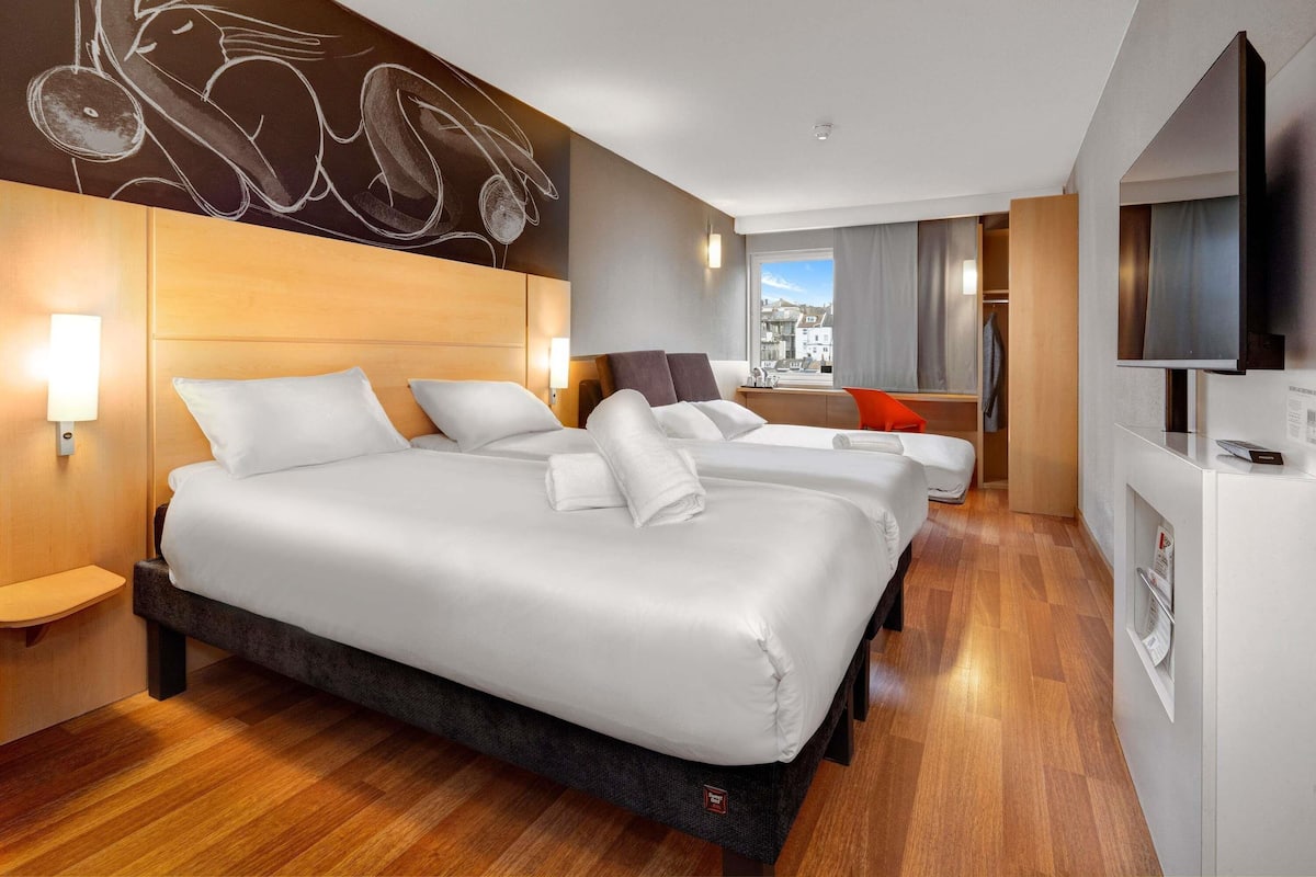 Habitación de hotel con tres camas, cabecera de madera, televisor de pantalla plana grande y vista a la ciudad.