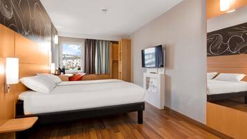 A hotel room with a large bed, a TV, a desk, and a window with curtains.