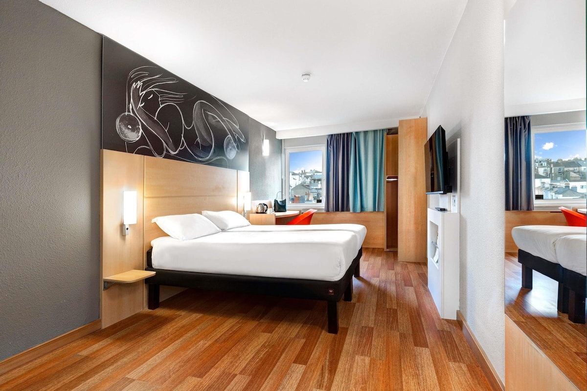 A modern hotel room with a bed, a desk, a chair, a TV, and a window with a city view.