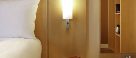 A neatly made bed with a white pillow, a bedside table with a lamp, and a small stack of books.