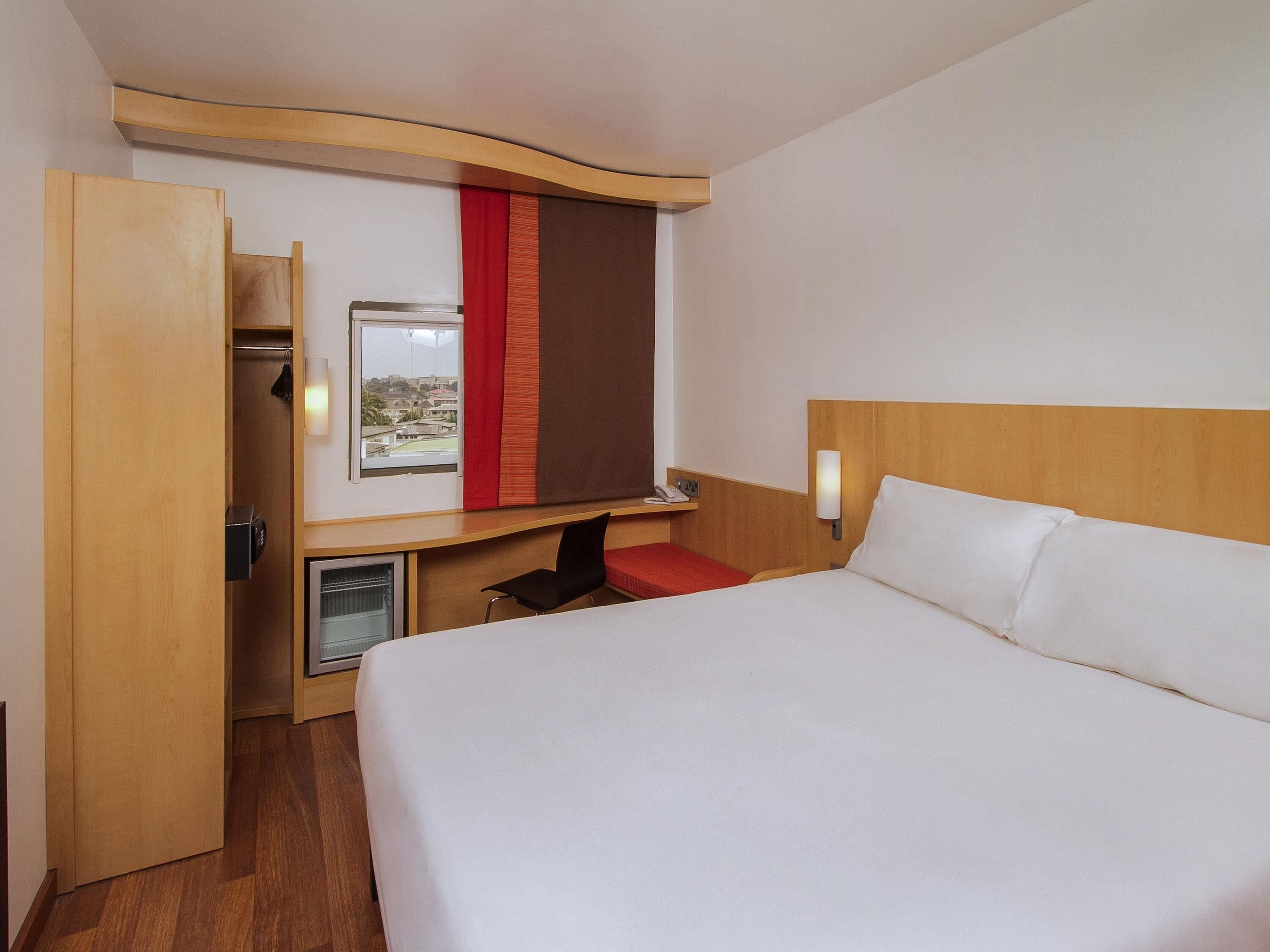 A hotel room with a bed, a desk with a chair, a small refrigerator, a telephone, and a window with a view.