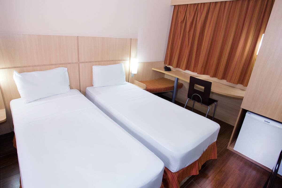 A hotel room with two single beds, a desk, a chair, and a small TV.