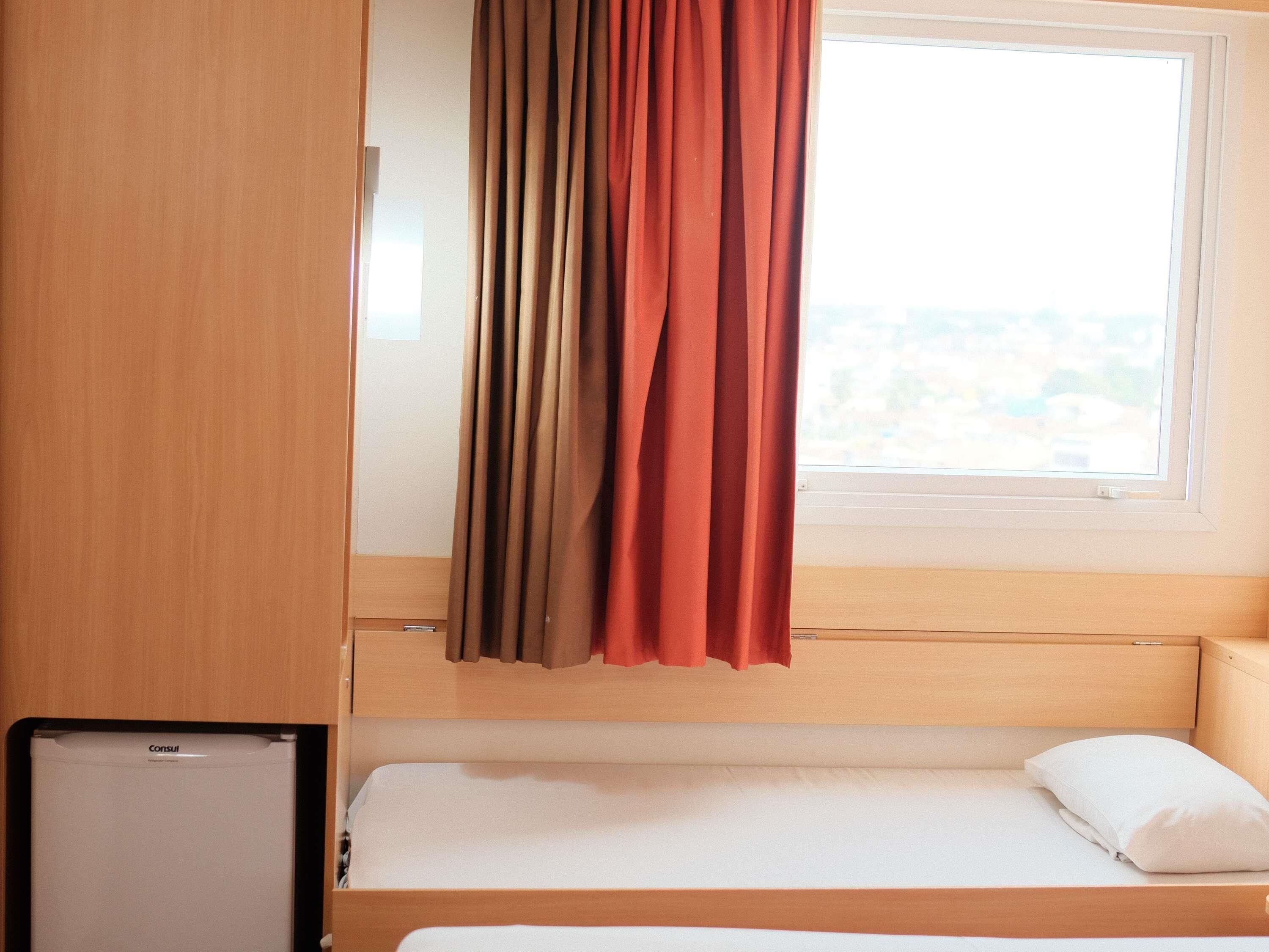 Una cama individual con almohada blanca, cabecera de madera, ventana con cortinas y una mesita de noche con un dispositivo.