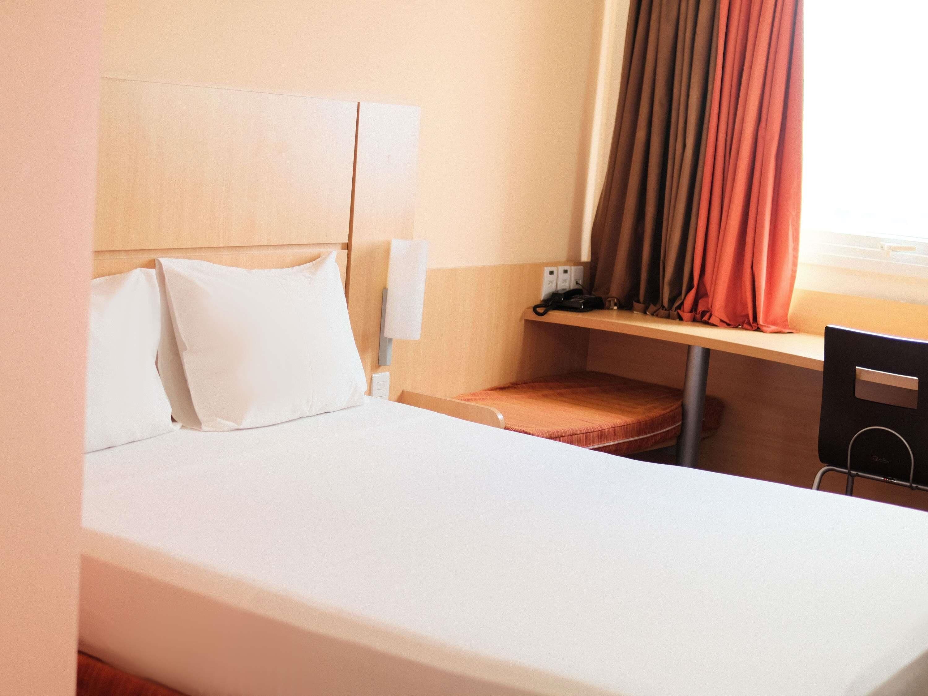 Une chambre d’hôtel avec un lit, un bureau, une chaise, une fenêtre avec des rideaux et une tête de lit.