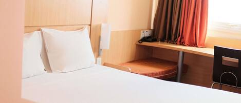 Habitación de hotel con cama, escritorio, silla, ventana con cortinas y cabecero.