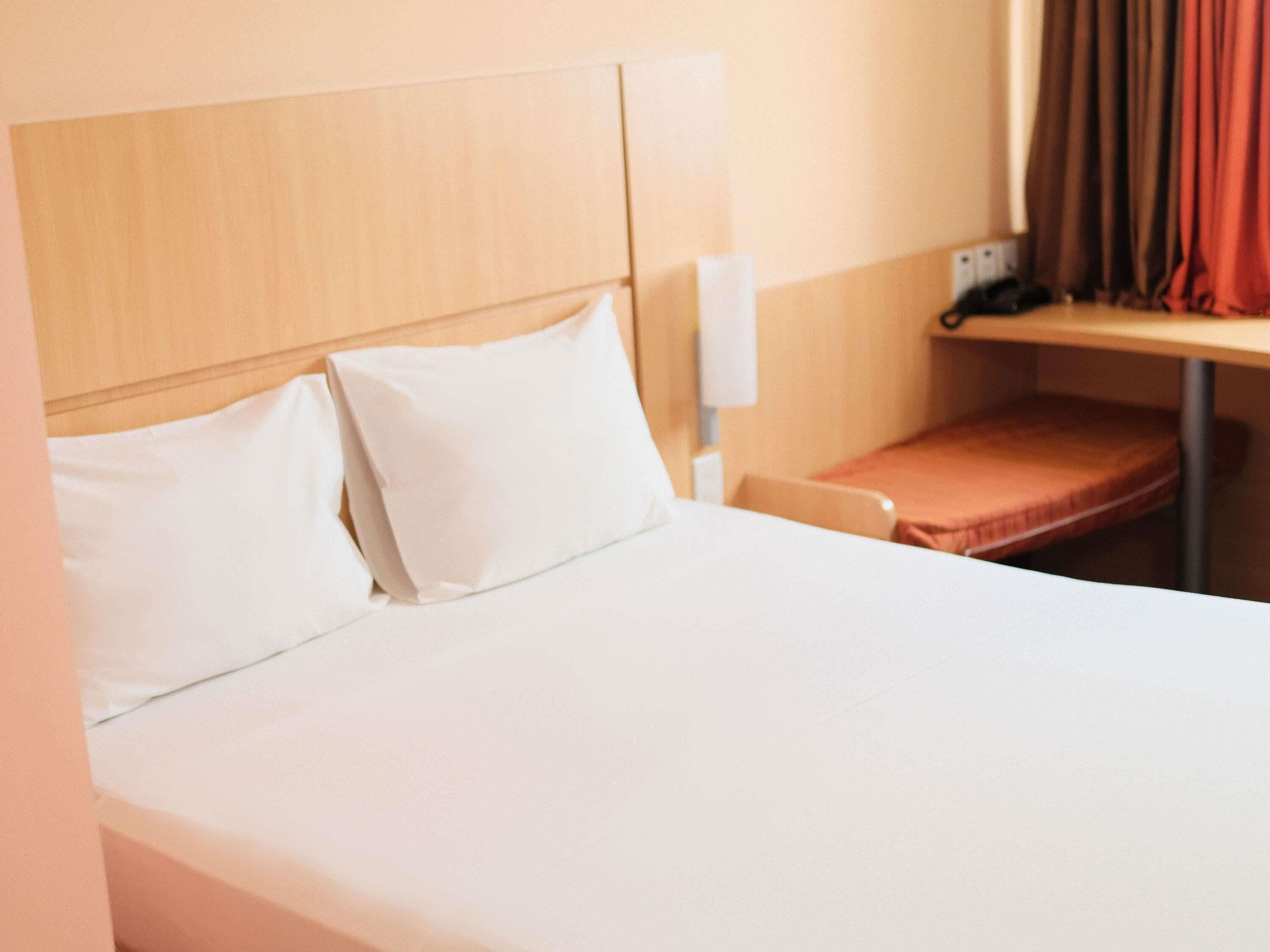 Quarto de hotel com cama, almofadas, mesa de cabeceira, telefone e janela com cortinas.