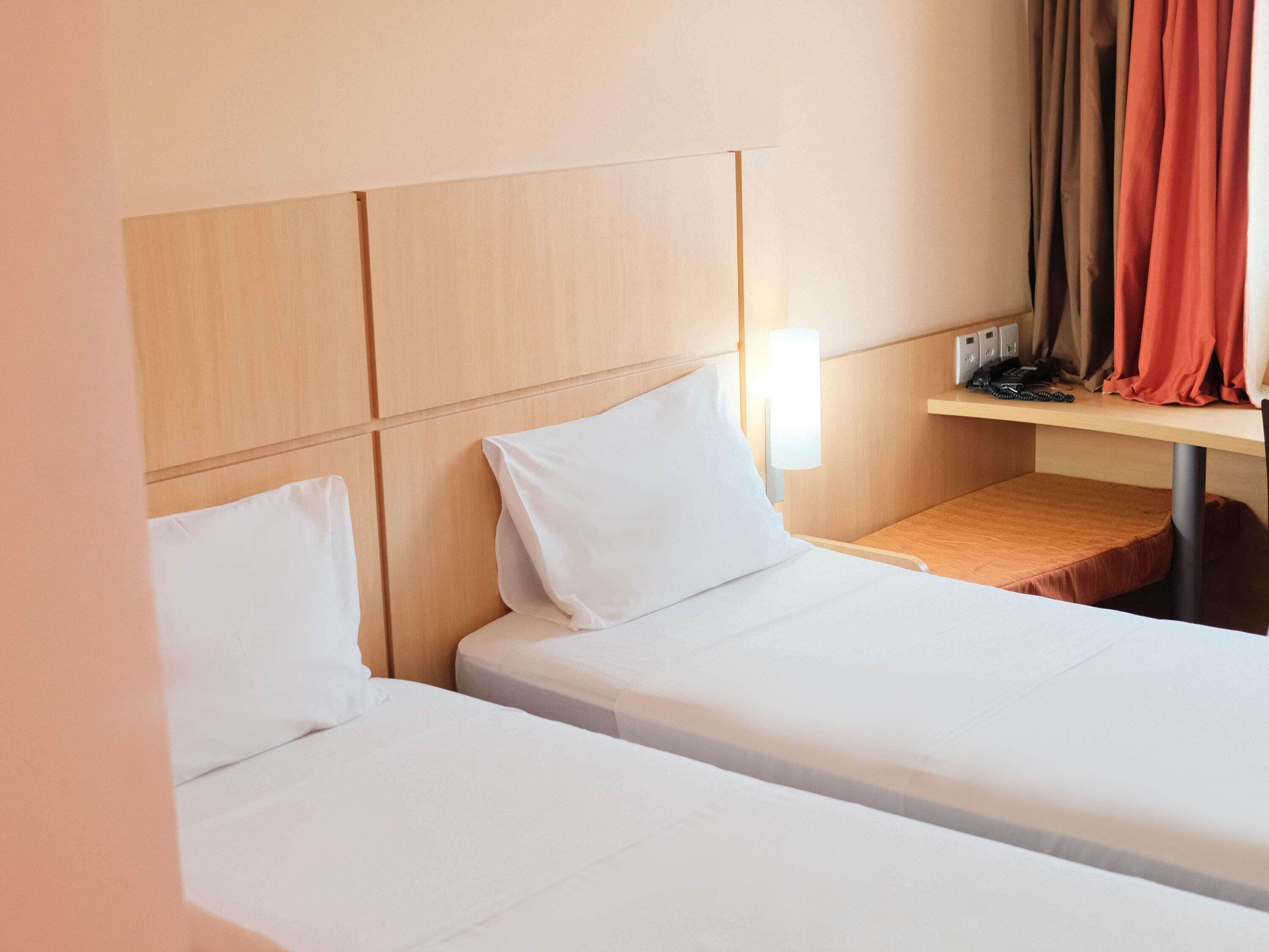 A hotel room with two beds, a bedside table, a lamp, and a window with curtains.