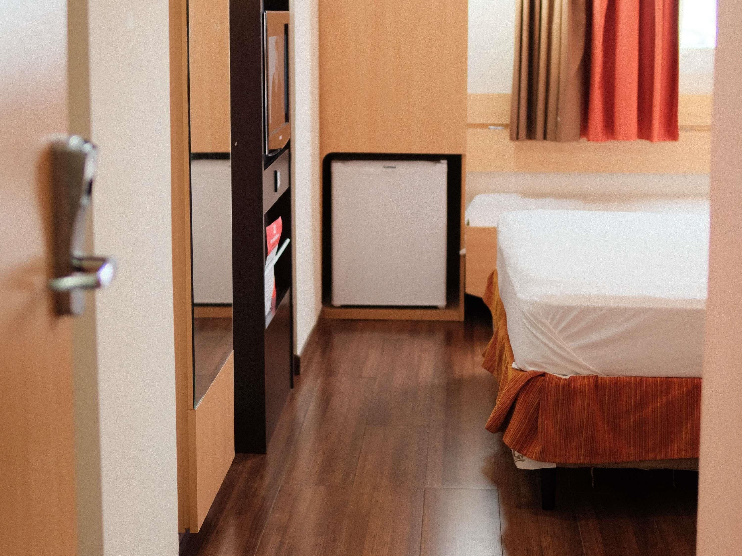 A narrow hallway with a bed on the right, a small refrigerator, and a door leading to a room.