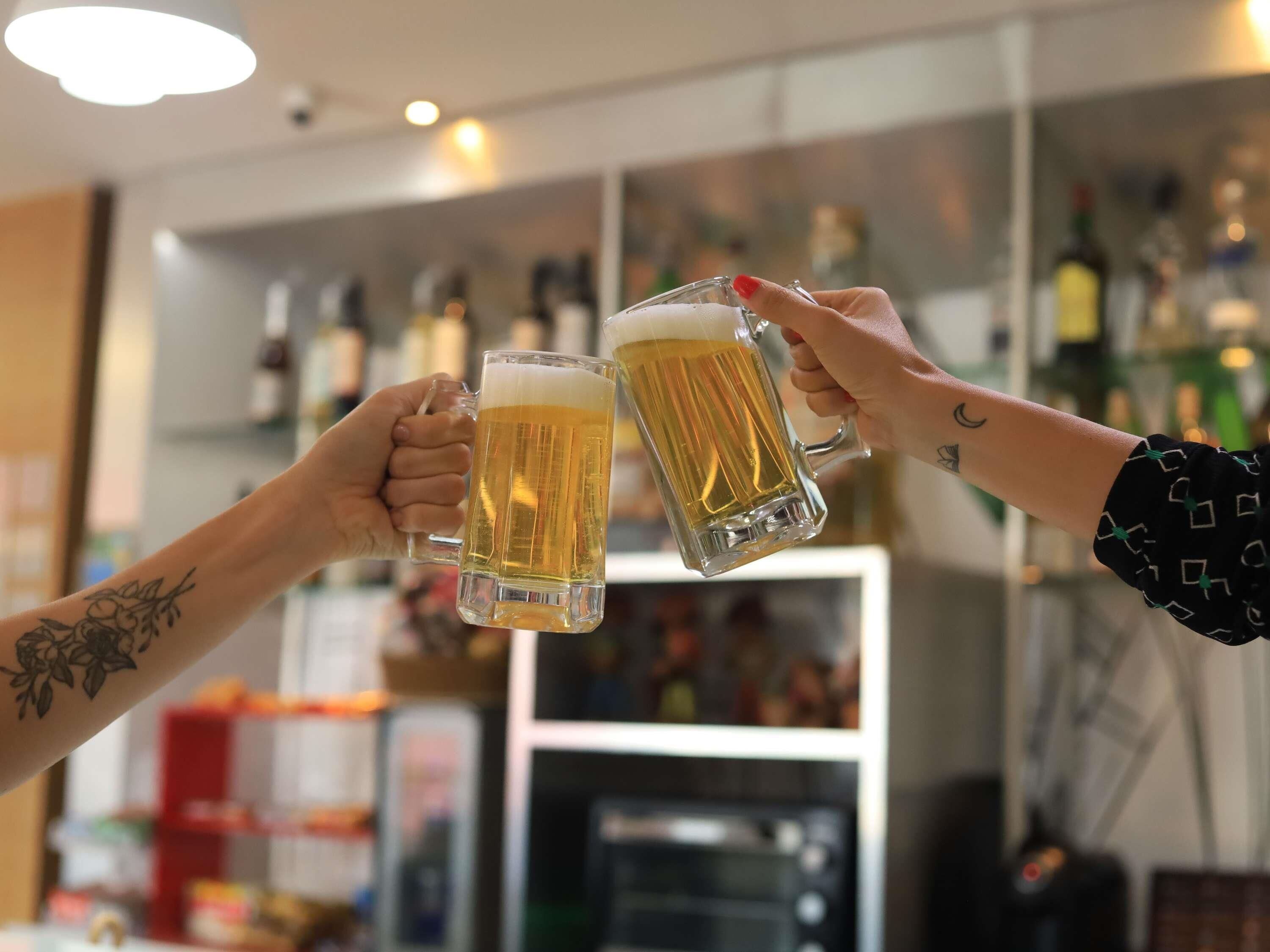 Two people toasting with beer mugs in a bar setting with shelves of bottles in the background.