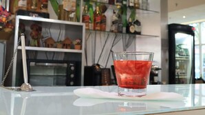 A bar area with a glass of red drink on a white napkin, shelves with various liquor bottles, and a decorative item on the counter. - Ibis Feira De Santana (Feira de Santana)