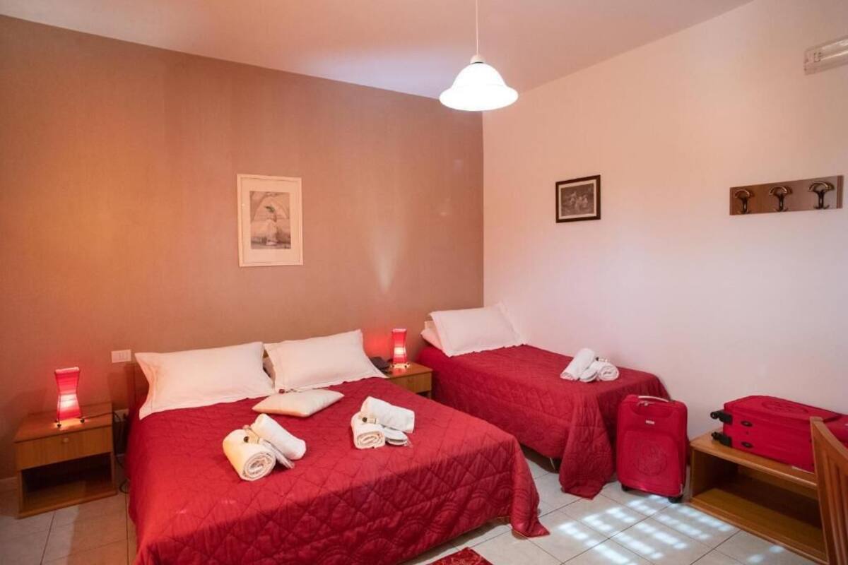 Habitación de hotel con dos camas, cada una con ropa de cama roja y almohadas blancas, una mesita de noche con una lámpara roja y una maleta roja en el suelo.