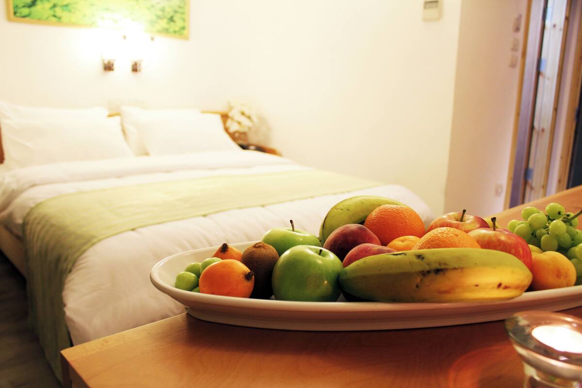 Un lit bien fait, recouvert d’une couette blanche et agrémenté d’oreillers, une table de chevet sur laquelle se trouve un panier de fruits contenant des pommes, des oranges et des bananes, et un petit verre d’eau.