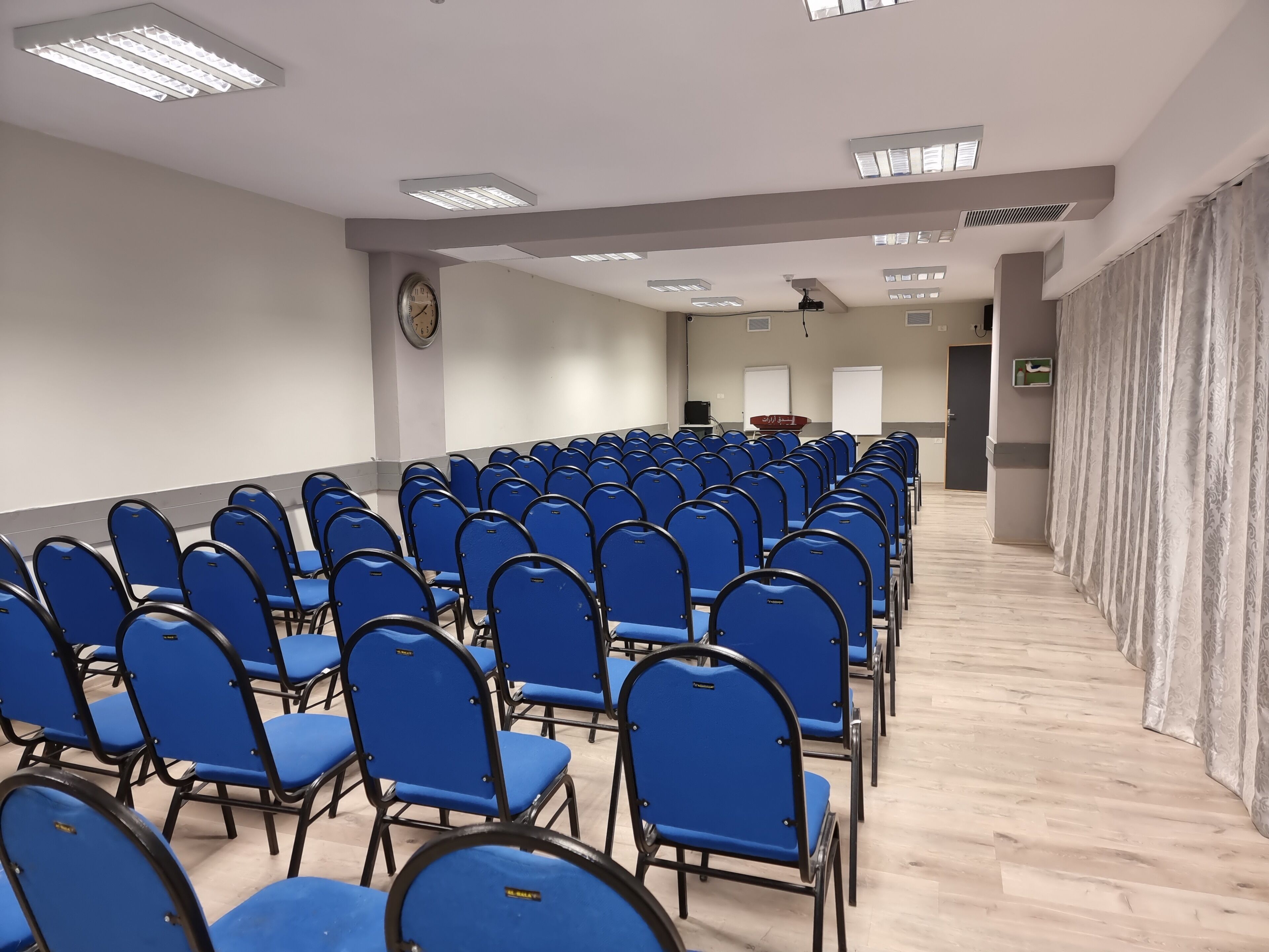 Un salón de conferencias con filas de sillas azules, un reloj en la pared y una pizarra al frente.