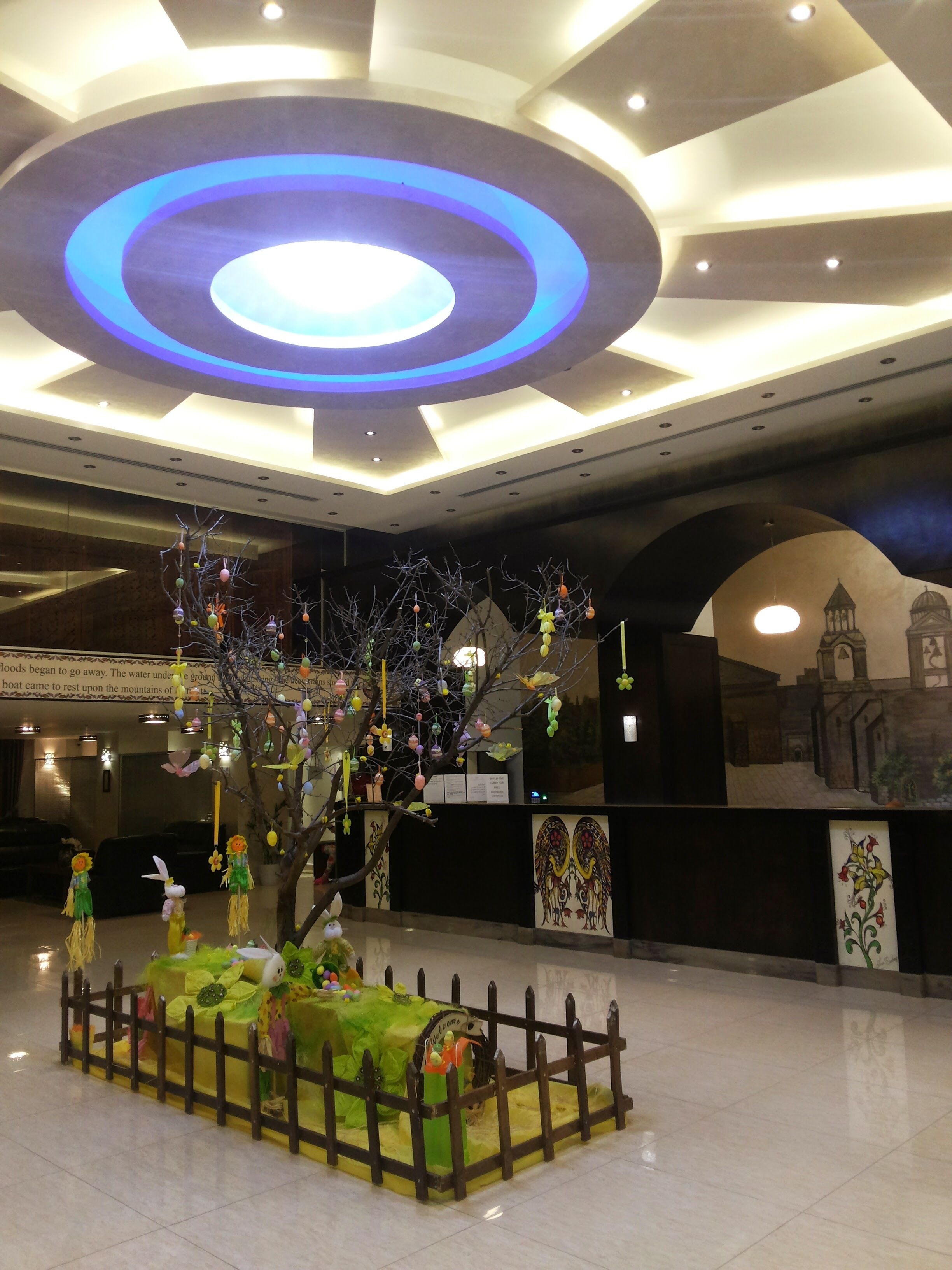 A lobby with a modern ceiling design, a decorative tree with ornaments, and a small fenced area with a bench.
