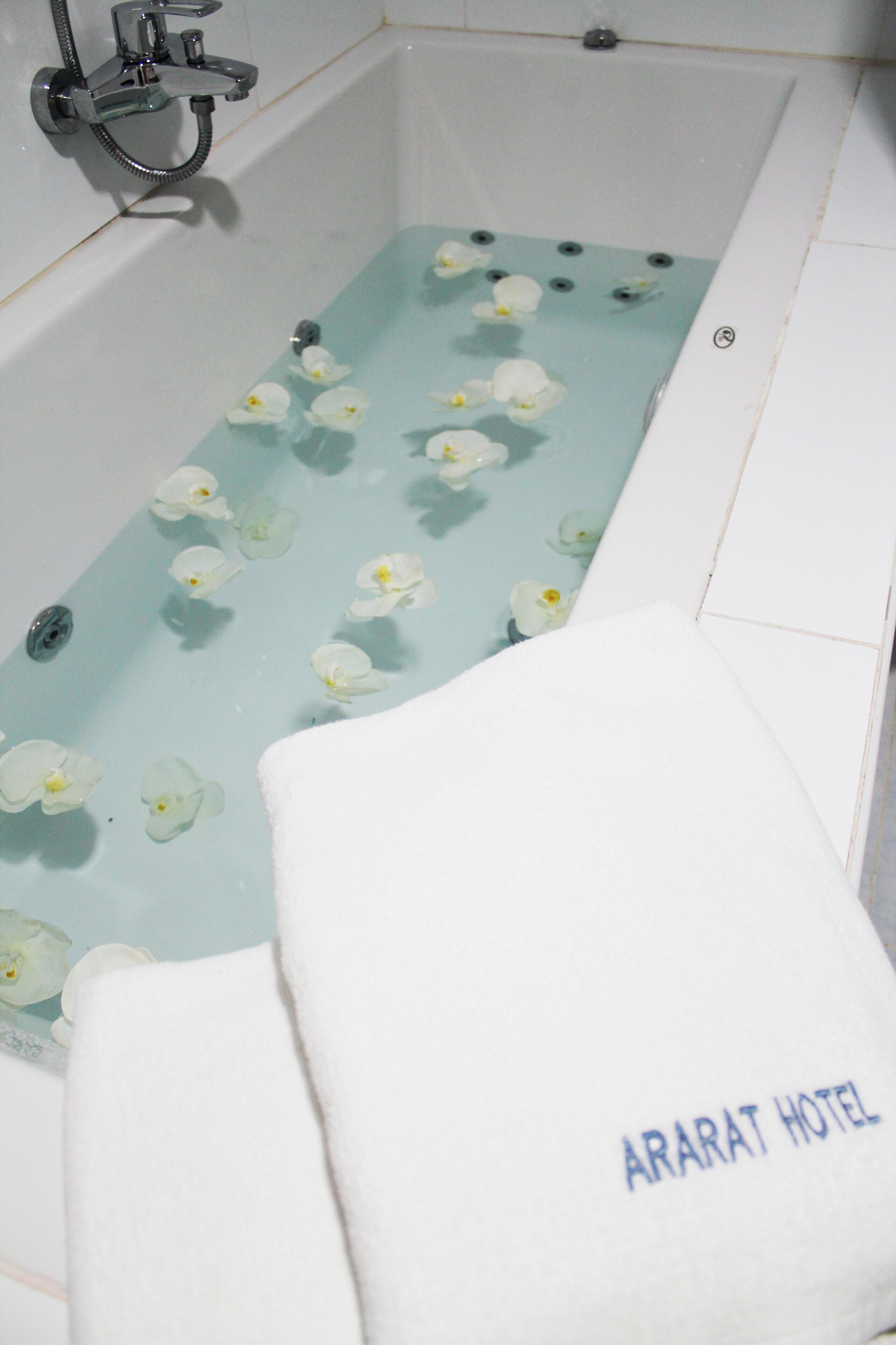 Una vasca da bagno con acqua e fiori bianchi, un soffione della doccia e un asciugamano con la scritta "ARARAT HOTEL".