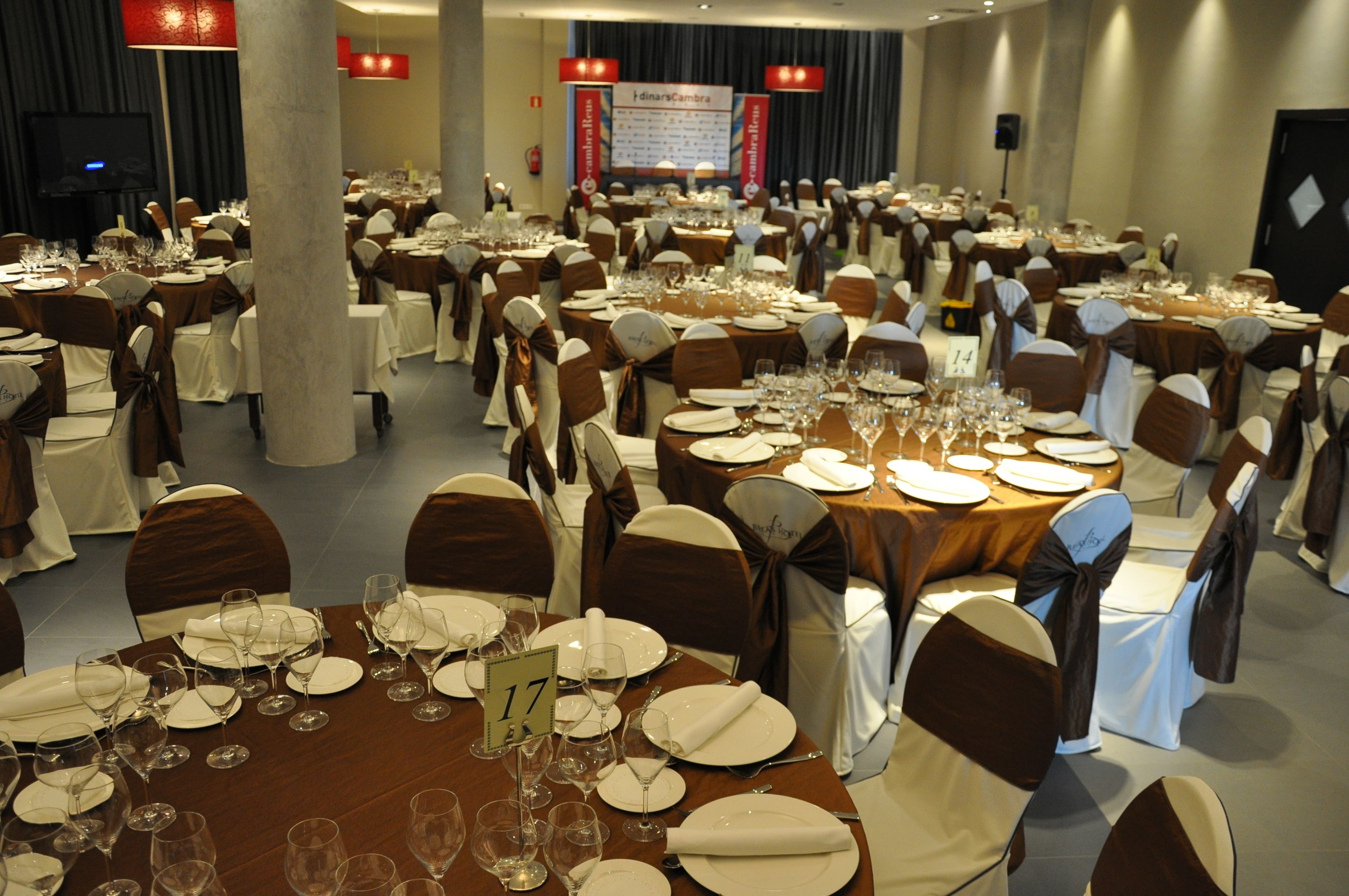 A banquet hall with round tables set for dining, numbered signs, and chairs with brown sashes.