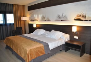 Una cama doble con colcha marrón y gris, dos mesitas de noche con lámparas, una silla y una pared con apliques de luz con forma de libros.