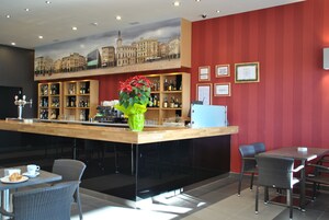 Zona de bar con mostrador de madera, estanterías con botellas y una planta decorativa. Hay sillas y mesas de mimbre, con una pared roja al fondo.