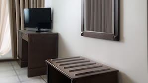 Habitación de hotel con televisión de pantalla plana sobre un escritorio de madera, un espejo grande en la pared y una ventana con cortinas color beige.