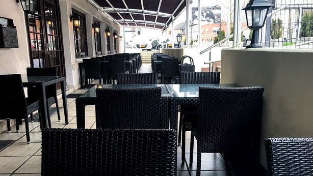 Un café al aire libre con mobiliario de mimbre negro, una zona cubierta para sentarse y vistas a la calle.