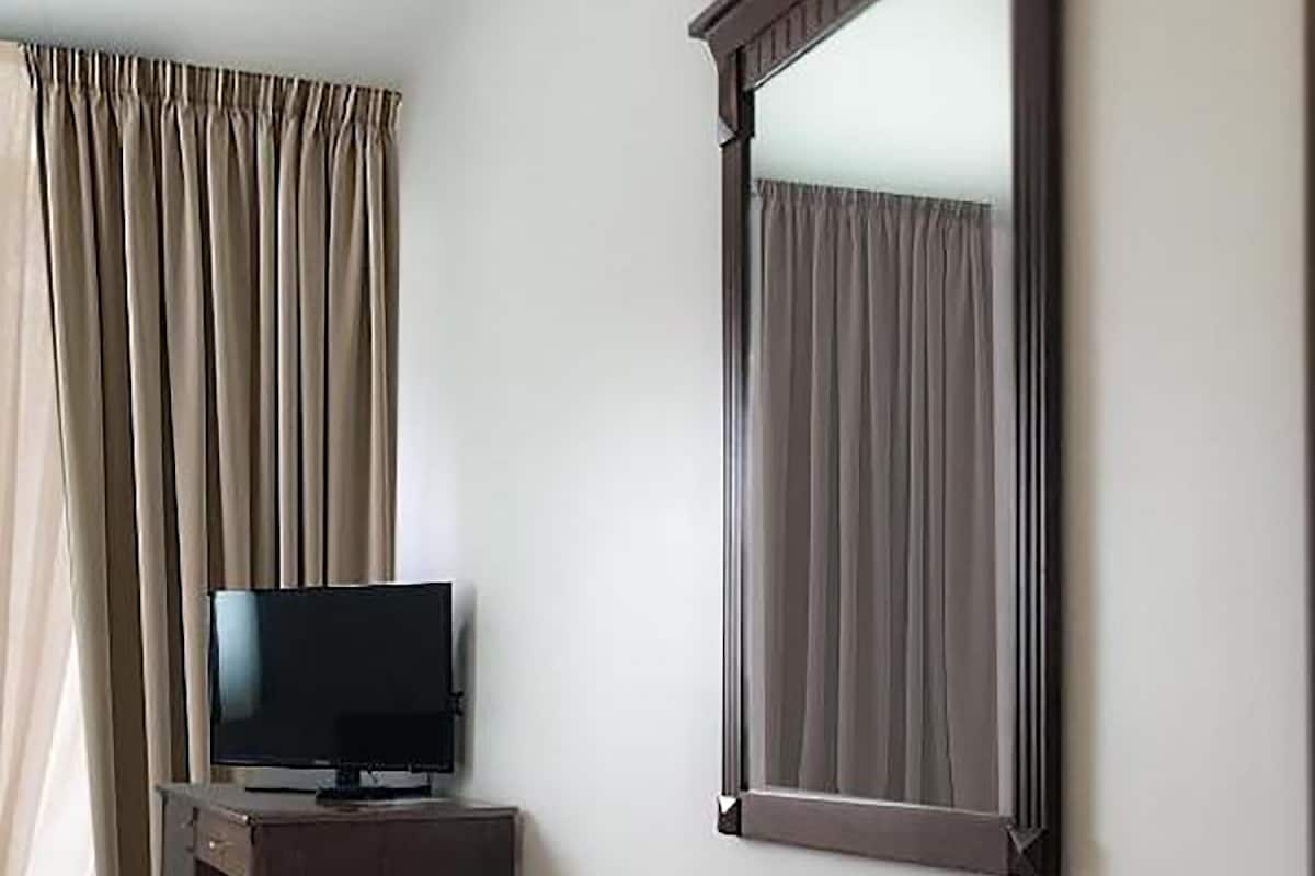 A hotel room with a flat-screen TV, a small wooden side table, a mirror, and beige curtains.