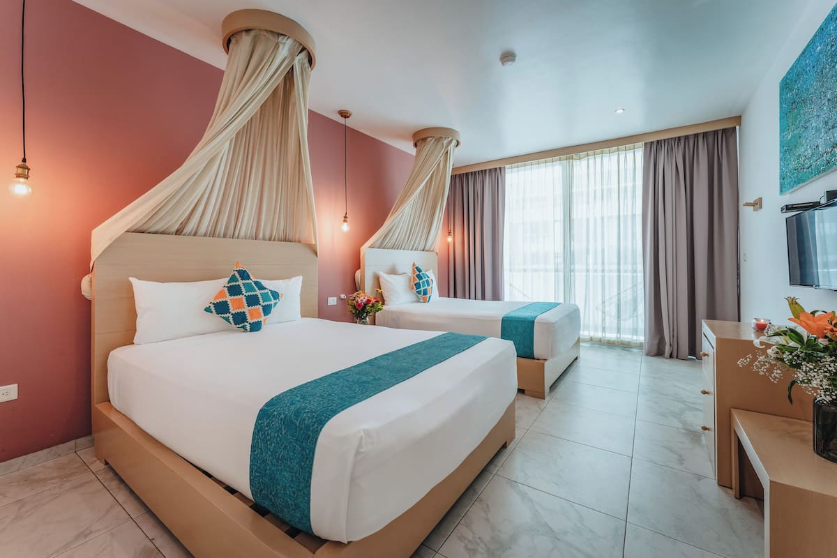 A hotel room with two beds, a TV, a desk, and a large window with curtains.
