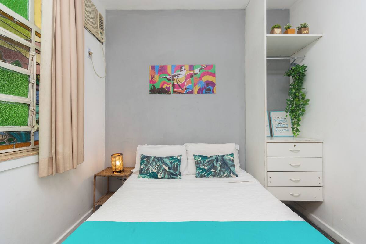 A bedroom with a bed, a nightstand, a wall-mounted shelf with plants, and a window with curtains.