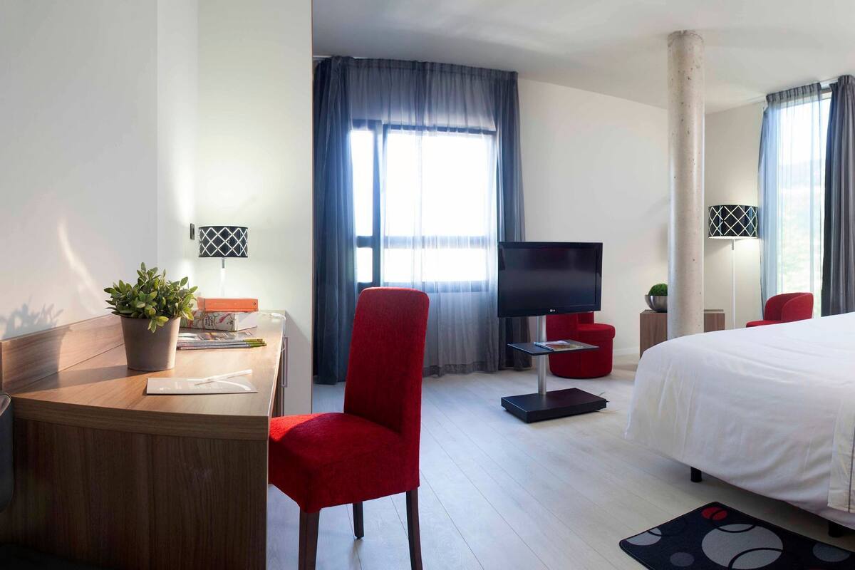 A hotel room with a bed, a desk with a chair, a TV, and a window with curtains.